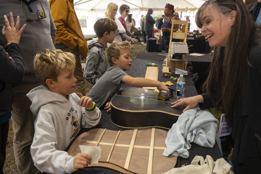 Three young boys look at instrument-maker Lisa Ring's guitar pieces at the 2022 Richmond Folk Festival. 