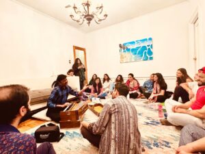A group of people, all sitting on the floor, gather together in a room tin front of one musician who is leading the group in singing and sitting in front of a harmonium.