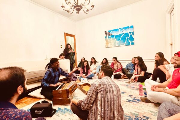 A group of people, all sitting on the floor, gather together in a room tin front of one musician who is leading the group in singing and sitting in front of a harmonium.
