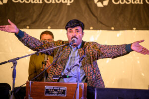 A man plays the harmonium with his hands spread wide and sings into a microphone.