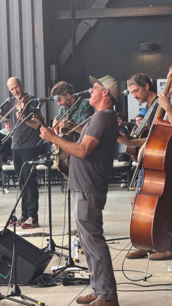 A group of musicians stands on a stage. In the foreground a man wearing black pants and a black shirt plays the mandolin.