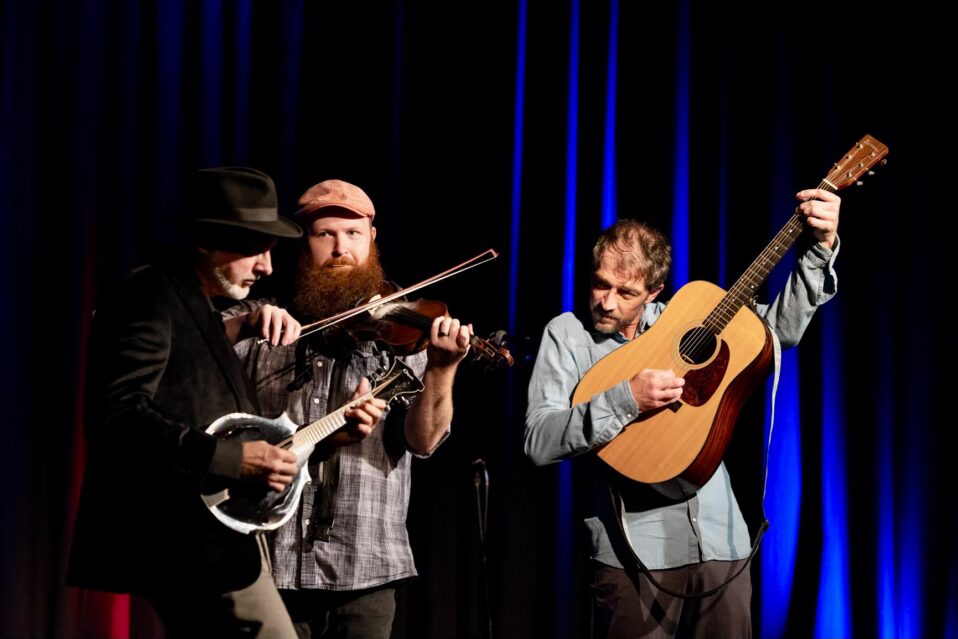 Three men stand on stage in front of a black curtain. The one on the left holds a mandolin, the one in the middle a fiddle, and the one on the right, a guitar.