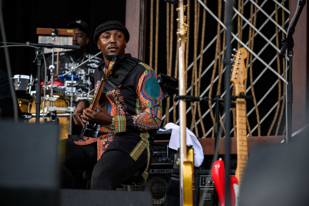 A man sits on a stool with a bass guitar in his hands and surrounded by other instruments.