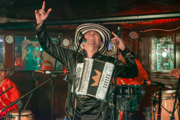 A man stands in front of microphone with his hands raised in the air. He has an accordion attached to his chest.