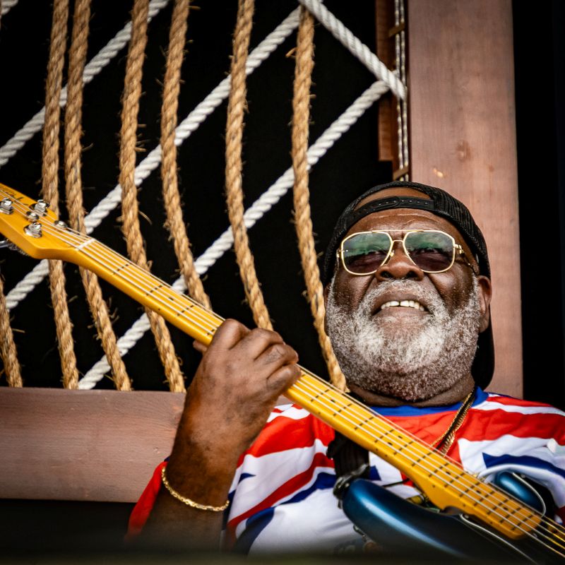 A man wearing sunglasses and a backwards baseball hat holds a bass guitar and smiles.