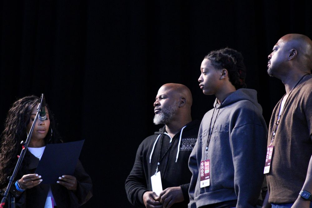 A woman stands in front of a microphone while three men stand to her left.