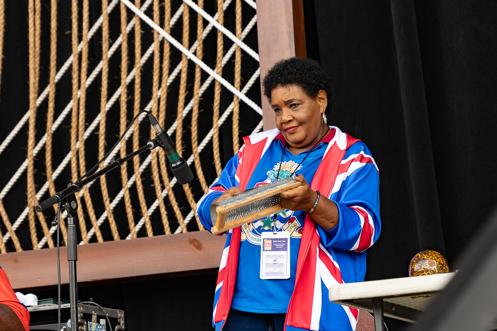 A woman with short heair wearing a colorful red, white, and blue jacket featuring the Caymanian flag holds a coconut grater in her left hand and a tool to scrape against it in her right to make a percussion nose.