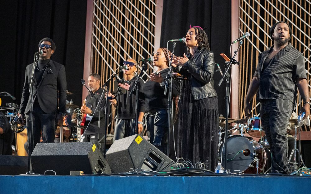 Singers and musicians wearing all black perform in front of black curtain on a stage.