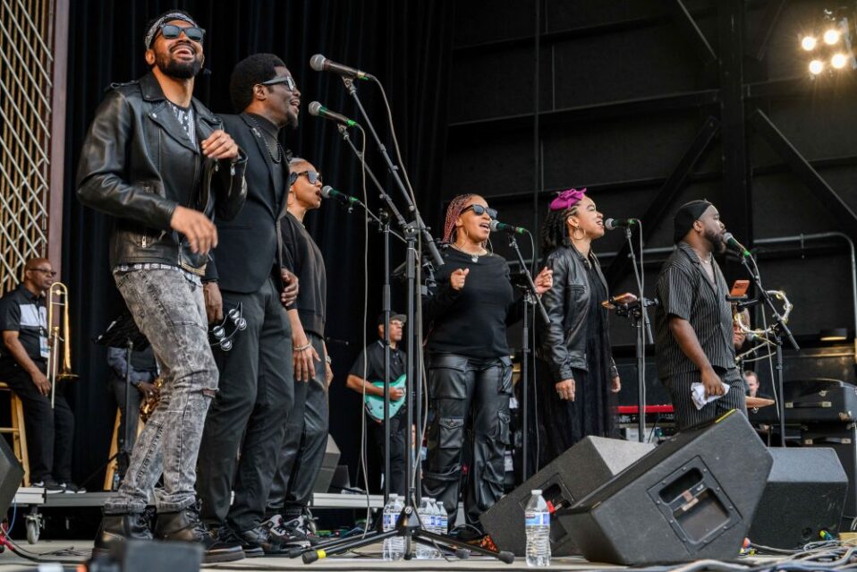 6 people wearing all black stand on stage in front of mirophones