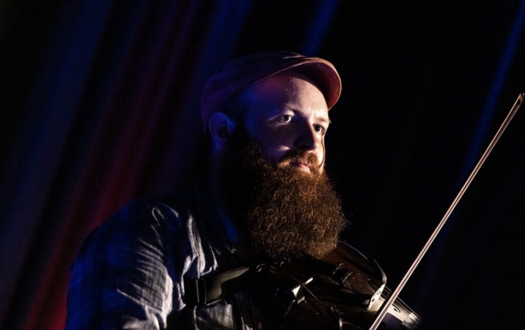 A closeup of a man with a bug beard and holding a fiddle.