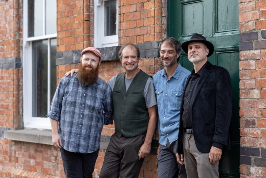 Four men stand in a row in front of a brick building.