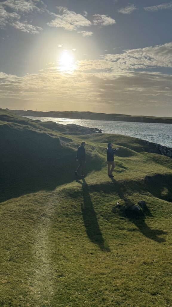 Two people walk along a green grassy hill overlooking blue water.